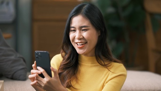 快乐微笑的亚洲年轻女子穿着黄色毛衣坐在沙发上，在家里用智能手机打视频电话