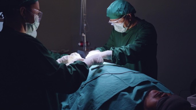 在黑暗的手术室里，一名护士从医疗托盘中取出镊子和聚丙烯线递给医生进行手术缝合，一名助手用棉垫吸血。