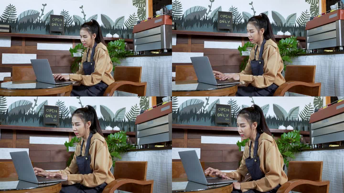穿着围裙的年轻亚洲咖啡店员工在桌子上摆弄笔记本电脑。咖啡厅工作人员下班后在电脑上查看新闻和更新在线内