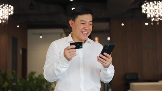 亚洲商人在通过个人智能手机成功实现信用卡在线支付时感到兴奋。穿白衬衫的先生待在办公室里，欢欣鼓舞。