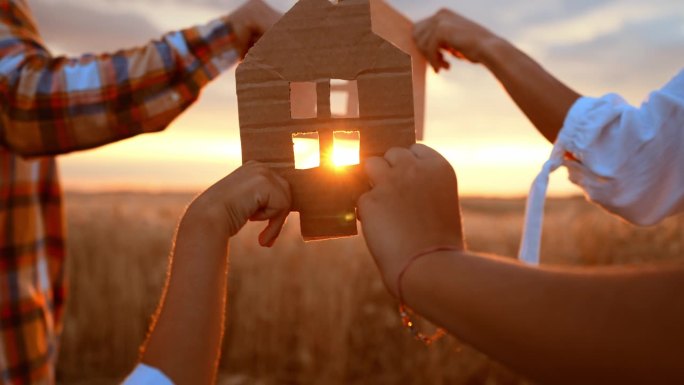 近距离用硬纸板做成的小模型纸房子，用手捧在麦田里，夏日的夕阳下。概念房屋贷款、抵押、拆迁、生态、建筑