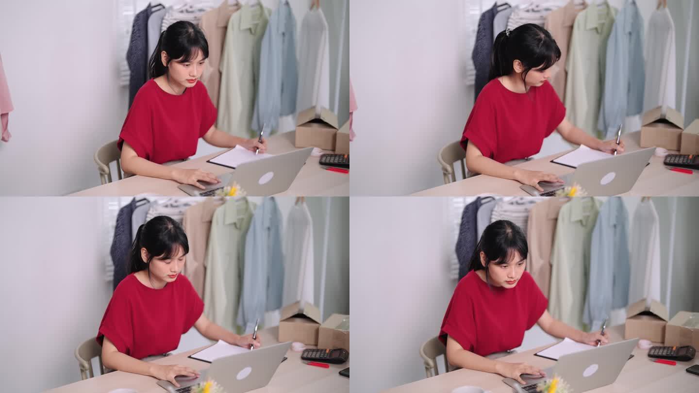亚洲在线服装店老板在笔记本电脑上勤奋地工作，全心全意地致力于卖衣服。捕捉她集中精力在她的家或商店设置