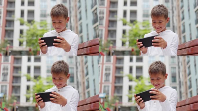 欢快的小家伙坐在外面的长凳上，全神贯注地玩着他的智能手机游戏，兴奋地敲击着键盘。小玩家在户外玩电子游