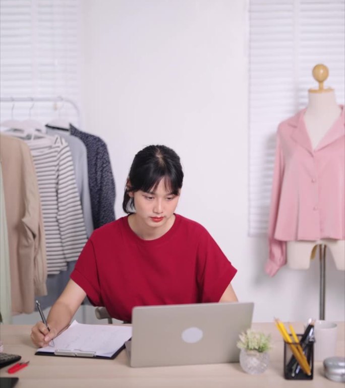 亚洲在线服装店老板在笔记本电脑上勤奋地工作，全心全意地致力于卖衣服。捕捉她集中精力在她的家或商店设置