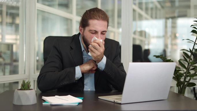办公室里生病的帅哥一边咳嗽一边用纸巾擤鼻涕，一边用现代笔记本电脑坐在桌子旁。我是一个时髦的白人商人，