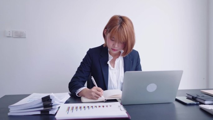 一位自信的亚洲女商人在她专门的工作空间里，孜孜不倦地把公司的重要数据从笔记本电脑记录到记事本上