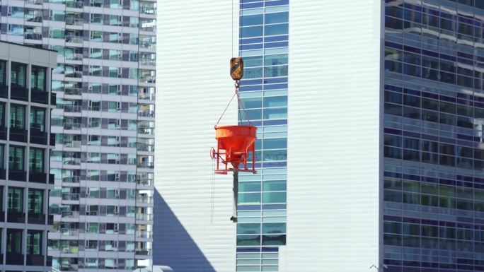 有窗户的新建筑前，起重机上吊着一个橙色的混凝土桶。用钩子吊着一桶水泥，上面画着一张脸。悬挂在空中的钢