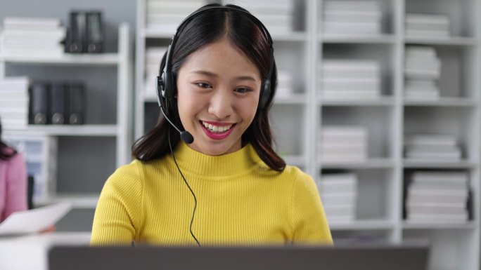 年轻的亚洲女性电话接线员，在办公室用电脑与客户交谈。