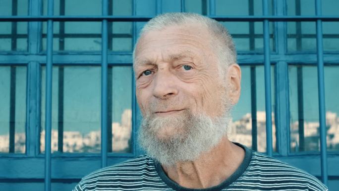 头部拍摄的慢动作肖像老人做微笑快乐的表情看着镜头。老年人的生活方式。白种成年男性，蓝眼睛，白胡子和白