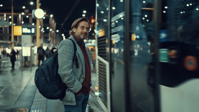 微笑的成熟商人背着双肩包站在路边，晚上公共汽车离开城市车站