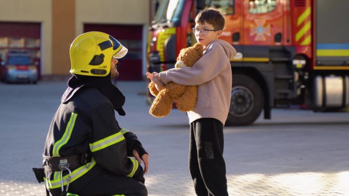 在消防站，小男孩拿着玩具和穿着防护服的消防员