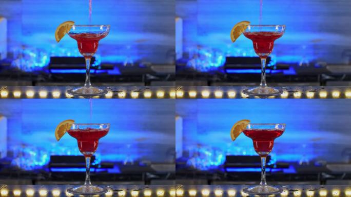 在蓝色背景灯光下的高档夜总会里，将一杯红色饮料倒入一杯橙色切片的鸡尾酒杯