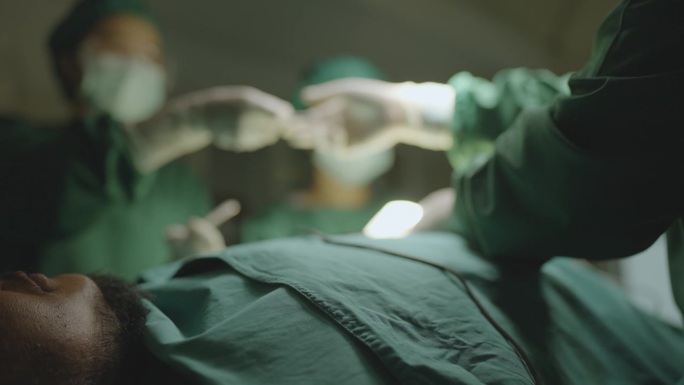 一名男性患者麻醉后躺在手术床上的特写，以及在低照度的手术室中，一名外科医生和他的团队在照明的手术灯下
