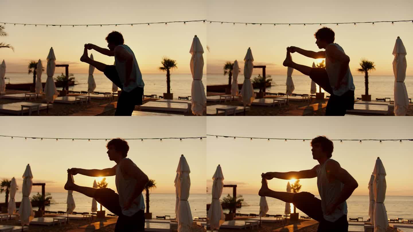 在一个阳光明媚的早晨，一个年轻人抬起腿伸了个懒腰。早上在阳光海滩做运动和瑜伽。健康的生活方式