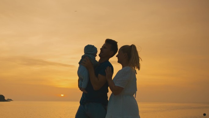 克罗地亚罗维尼，夕阳西下，一对快乐的父母带着刚出生的婴儿在海边橙色的天空下玩耍