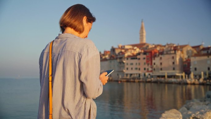 清晨漫步:女人在罗维尼日出的宁静美景中，在码头上用手机捕捉老城的魅力