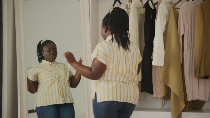 黑人女性减肥后在镜子前欣赏自己。快乐的非洲超重女性在家里跳舞。加码女性测量腰围。
