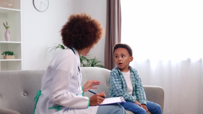非裔美国儿科医生坐在沙发上，和一个学龄前的小男孩在家里体检时聊得很开心。好心的医生在膝盖上用写字板记