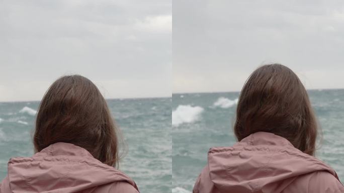 垂直视频。一个孤独的女人凝视着远处汹涌的大海，海浪冲击着尖锐的岩石。缓慢的运动。