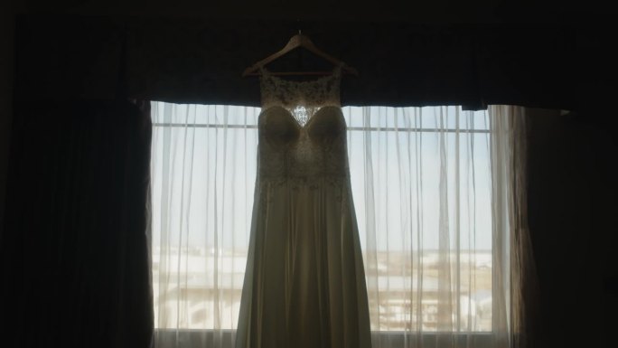 美丽的白色婚纱挂在衣架上，背光窗帘，在电影的慢镜头中，喜怒无常的风景在移动。