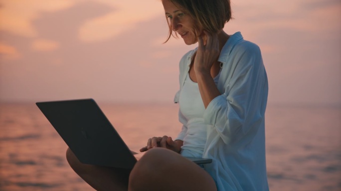 SLO MO海滨工作空间:孤独的女人坐在码头上用笔记本电脑工作，夜幕降临