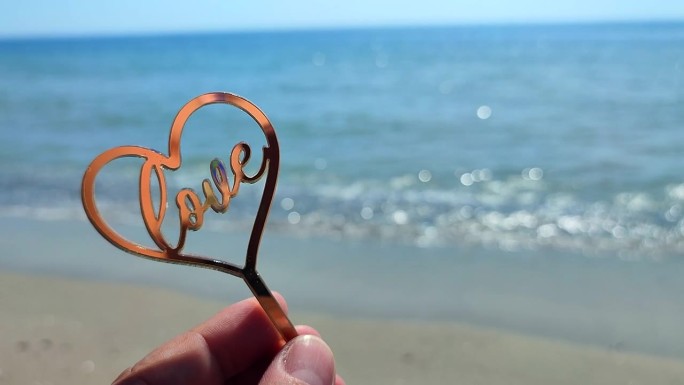 人拿在手指，手棍形状金色的心和字爱的背景海和海浪在海边在阳光明媚的夏日的特写。概念爱情浪漫爱情情人节