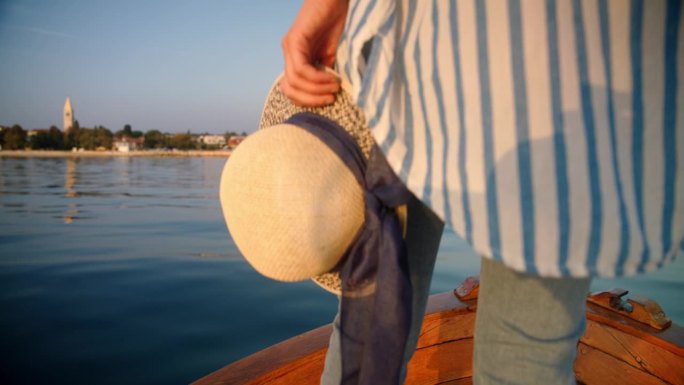 一个不知名的女人站在船上，拿着一顶帽子，在黄昏驶向岛屿