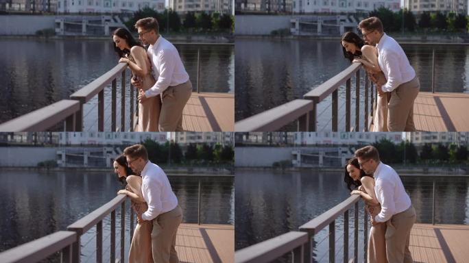 慢镜头，侧面视图。一对夫妇在城市堤岸的围栏码头上。一个女人透过障碍物向外看，一个男人从后面试图把她扶