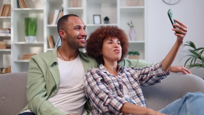 在现代智能手机上进行视频通话时，快乐的非裔美国夫妇互相倚靠。一对热恋的情侣躺在灰色的沙发上，在网上和