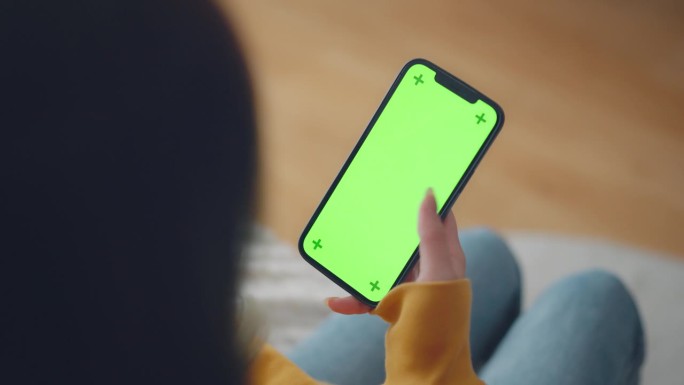 女人手拿着手机，坐在家里的沙发上，拿着智能手机的色度键模型，用空白的绿色屏幕滑动照片或图片