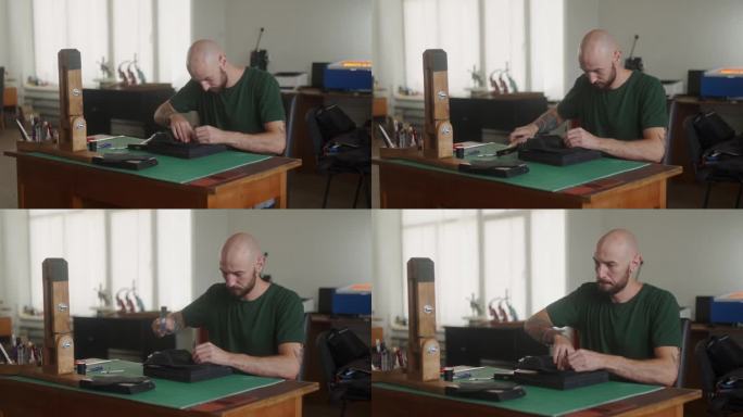 专业的皮革工匠在工作台上用皮革打头和锤子在黑色手袋上缝洞。穿着卡其布t恤的大胡子男子在车间缝钱包