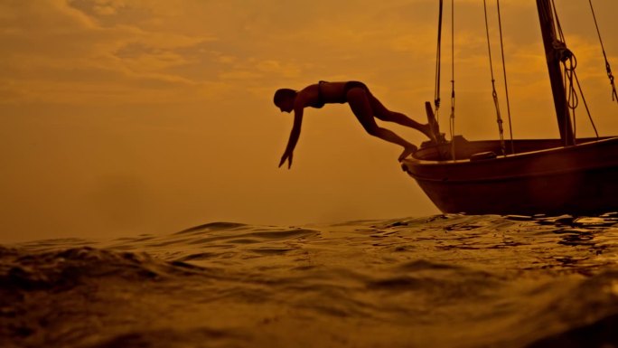 SLO MO黄昏潜水:日落时分，女人从船上跳入大海，玩得很开心