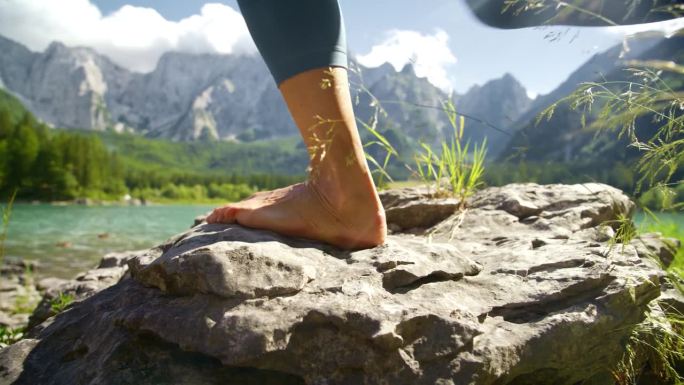 赤脚的女人踩在阳光明媚的湖边的岩石上