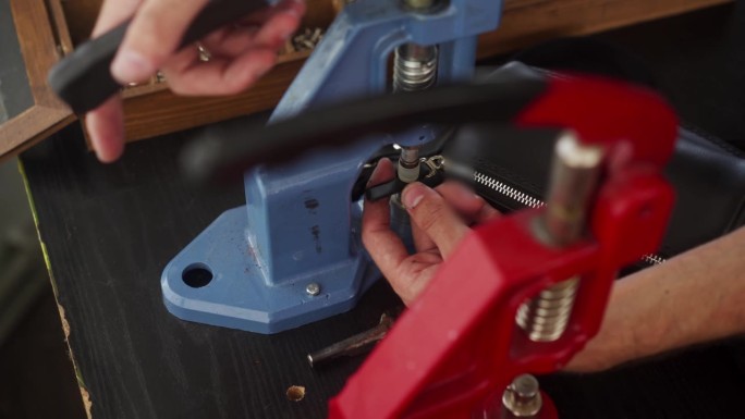 专业皮革工匠用手压机将别针固定在手提包的拉链上，用于安装金属配件。有纹身的男子在车间用特殊工具制作钱