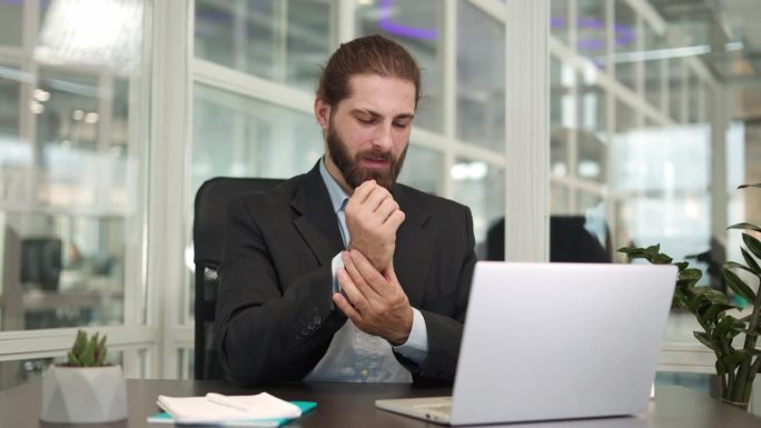 一名穿着时髦职业装的沮丧白人男子坐在办公桌旁，因为使用电脑而感到疼痛，他抚摸着手腕。因职业病导致手痛