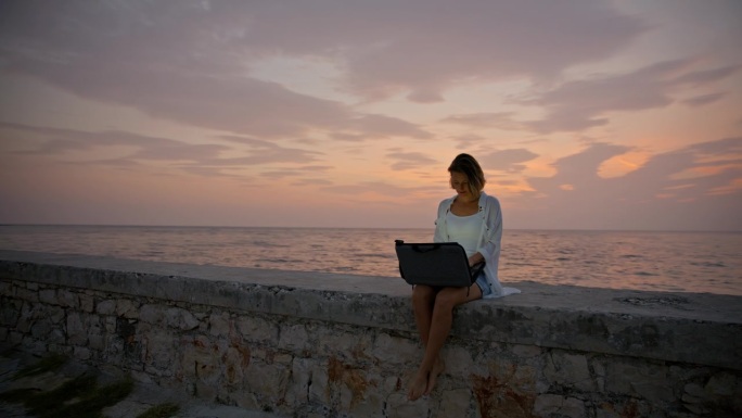 SLO MO海滨工作空间:黄昏时分，一名女子坐在码头上，用笔记本电脑工作