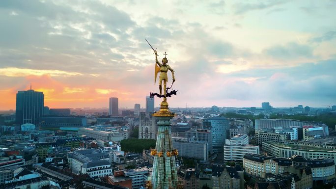 在日落时分，无人机在比利时布鲁塞尔市政厅塔尖上拍摄圣迈克尔雕像。背景上有多个建筑