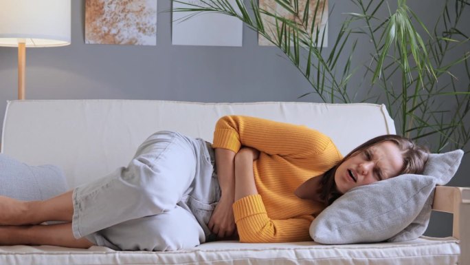 穿着黄色衬衫和牛仔裤的女人躺在客厅的沙发上，手放在肚子上，患有月经疼痛，痉挛和胃痛，患有慢性胃炎。