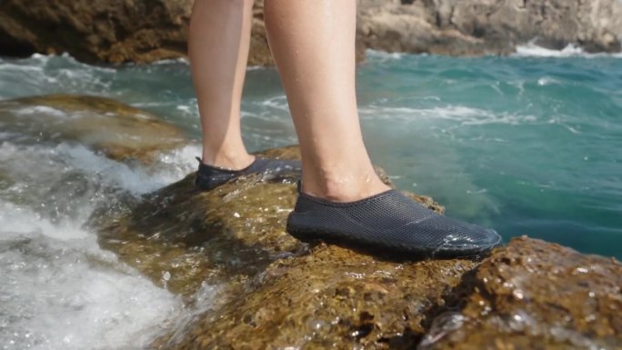 女性脚的特写镜头，她穿着专门为陡峭岩石设计的水鞋，站在岩石悬崖上，波浪覆盖着她的脚。缓慢的运动。