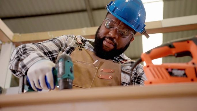 穿着防护服的非洲木匠在建造新家具前用电钻在木板上工作。在木工车间制造精工产品的专业工人。