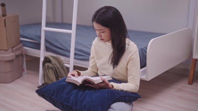 一名年轻的亚洲学生穿着休闲服坐在宿舍的双层床上，全神贯注地看书。在舒适的宿舍里，她靠在双层床凸起的靠