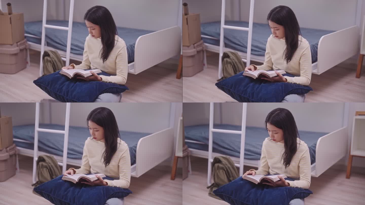一名年轻的亚洲学生穿着休闲服坐在宿舍的双层床上，全神贯注地看书。在舒适的宿舍里，她靠在双层床凸起的靠