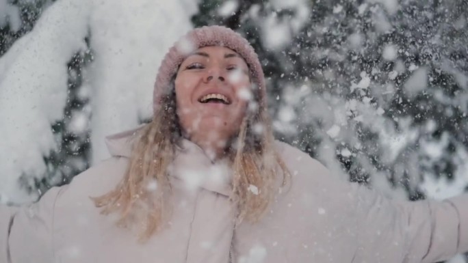 年轻女子对着镜头吹雪花。微笑的漂亮女孩，穿着白色的冬季夹克，戴着蓝色的针织帽，吹着手掌上的雪。快乐的