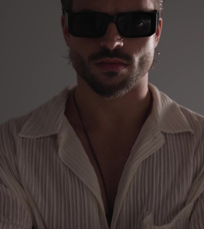 性感的时尚男人戴着太阳镜，双臂折叠，看向一侧和向前，在灰色背景上性感地触摸嘴唇