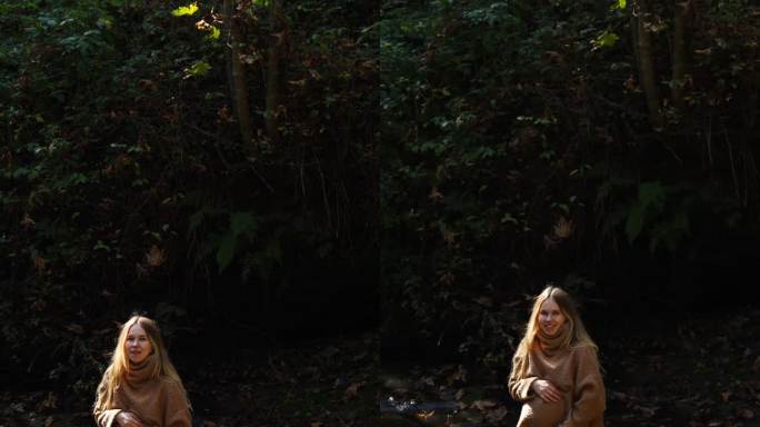 在一个阳光明媚的日子里，一位穿着毛衣的美丽孕妇站在山间河流背景的石头上，抚摸着自己的肚子，看着镜头。