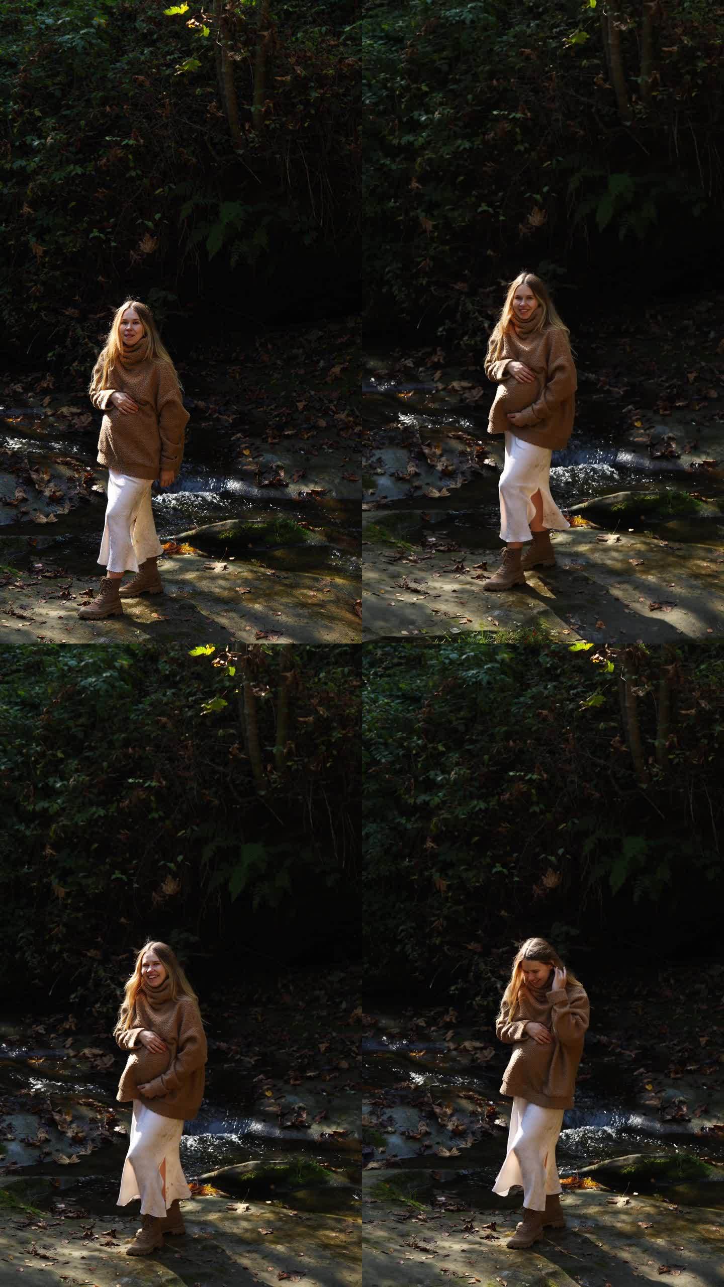 在一个阳光明媚的日子里，一位穿着毛衣的美丽孕妇站在山间河流背景的石头上，抚摸着自己的肚子，看着镜头。