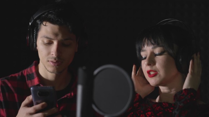 一个男人和一个女人，有抱负的艺术家，站在录音棚里唱一首歌。这名男子看着智能手机屏幕上的歌词。