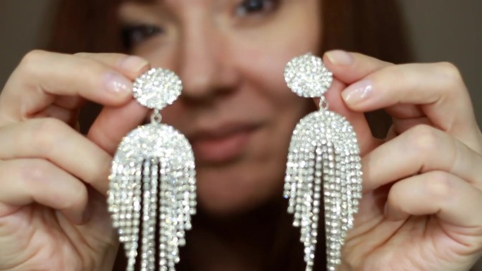 一个戴着大耳环的女人的特写。女孩戴着奢华昂贵的珠宝，上面有闪闪发光的钻石和宝石。女性双手紧握，露出耳