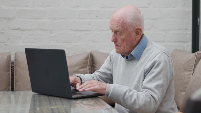 笔记本电脑，搜索和老人在沙发上进行网上购物，滚动或轮椅研究在他的家里。电脑，打字和残疾人在客厅进行电