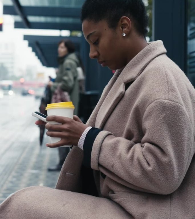 在城市的公交车站，穿着长外套的年轻女子一边喝咖啡一边玩手机
